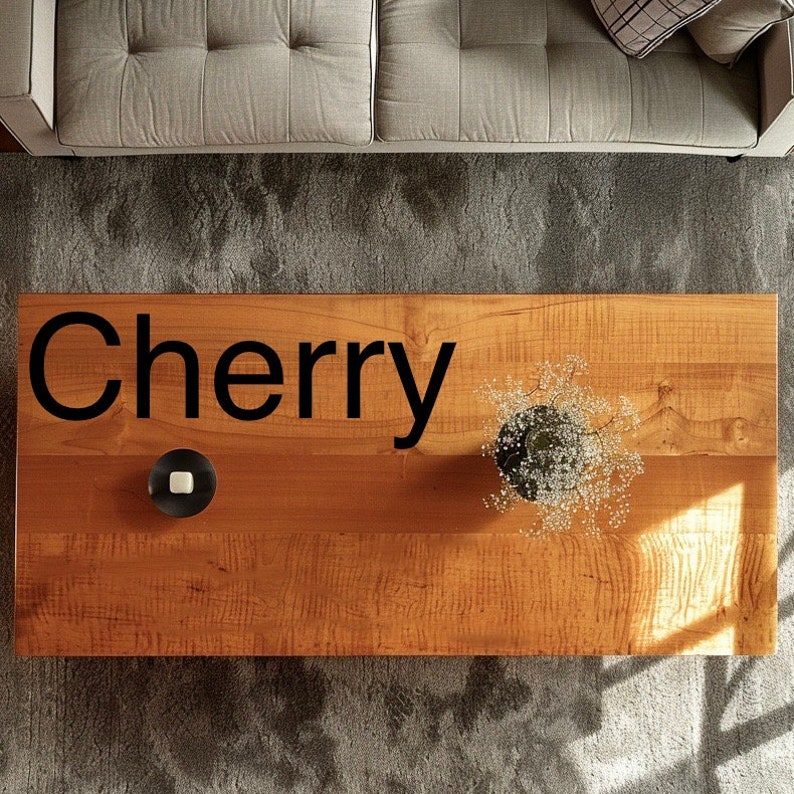 May include: A close-up of a wooden table top with the word "Cherry" written in black lettering. The table top is light brown with a natural wood grain pattern. There is a small white candle in a black holder and a small bouquet of white flowers in a black vase on the table.