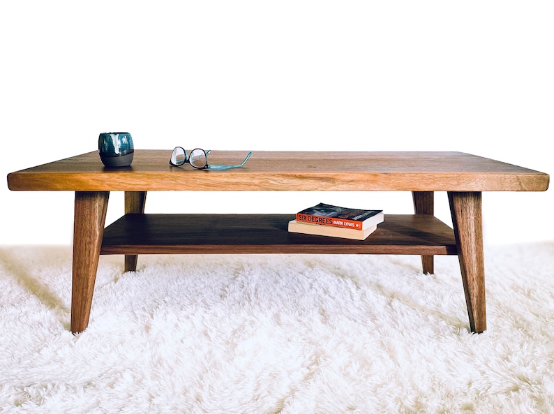 May include: A wooden coffee table with a lower shelf. The table has a natural wood finish and is sitting on a white shag rug. There are glasses and a book titled "Six Degrees" on the table.