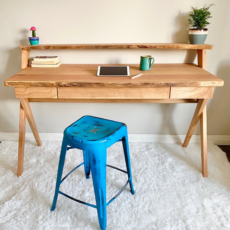 Pu&ograve; includere: Una scrivania in legno chiaro con piano a bordo vivo e ripiano, con cassetto e gambe angolate. Davanti c'&egrave; uno sgabello in metallo blu. La scrivania &egrave; accessoriata con un tablet, una tazza e piccole piante in vaso.