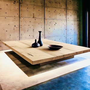 May include: A large, rectangular wooden table with a natural finish sits on a light brown rug in front of a concrete wall. Three black ceramic vases are arranged on the table, along with a brown bowl.