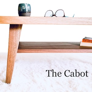 May include: A wooden coffee table with a lower shelf. The table has a light wood finish and angled legs. A book titled "Six Degrees" by Mark Lynas is on the shelf. The table is on a white fluffy rug. The text "The Cabot" is below the table.