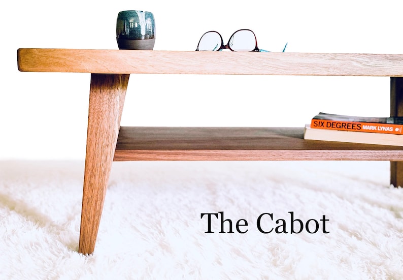 May include: A wooden coffee table with a lower shelf. The table has a light wood finish and angled legs. A book titled "Six Degrees" by Mark Lynas is on the shelf. The table is on a white fluffy rug. The text "The Cabot" is below the table.