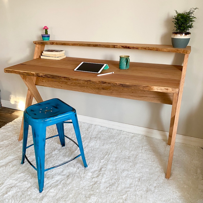 Pu&ograve; includere: Una scrivania in legno con un design a bordo vivo, con un ripiano integrato. Uno sgabello in metallo blu &egrave; posto di fronte alla scrivania. Un tablet, una tazza e piccole piante sono sulla scrivania. La scrivania &egrave; in legno chiaro.