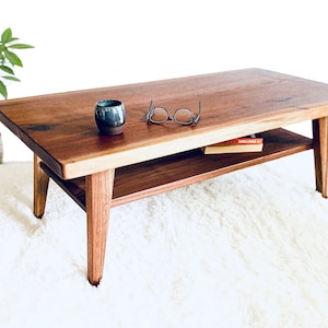 May include: A wooden coffee table with a lower shelf. The table has a natural wood finish and is sitting on a white rug. There are glasses and a book on the table.