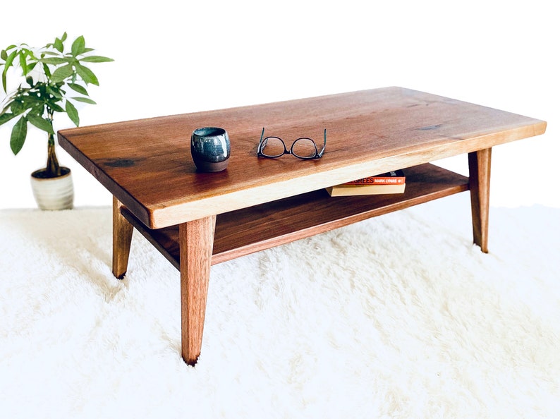 May include: A wooden coffee table with a lower shelf. The table has a natural wood finish and is sitting on a white rug. There are glasses and a book on the table.