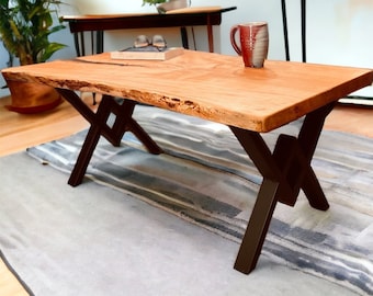 May include: A wooden coffee table with a live edge top and black metal X-shaped legs. The table is sitting on a gray and white striped rug.