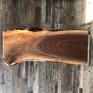 Pu&ograve; includere: Una lastra di legno rettangolare con bordo vivo, dalla forma naturale e organica. La venatura del legno presenta un ricco colore marrone scuro con riflessi marrone dorato pi&ugrave; chiari. La lastra &egrave; probabilmente destinata all'uso come tavolo o piano di lavoro.