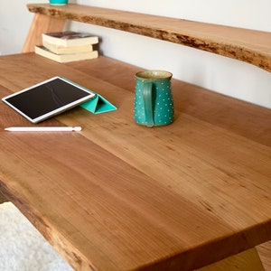 Pu&ograve; includere: Una scrivania in legno con bordo naturale e ripiano abbinato. Una tazza turchese con pois bianchi &egrave; appoggiata sulla scrivania, insieme a un tablet e uno stilo. Un piccolo cactus in un vaso blu e viola &egrave; sullo scaffale.