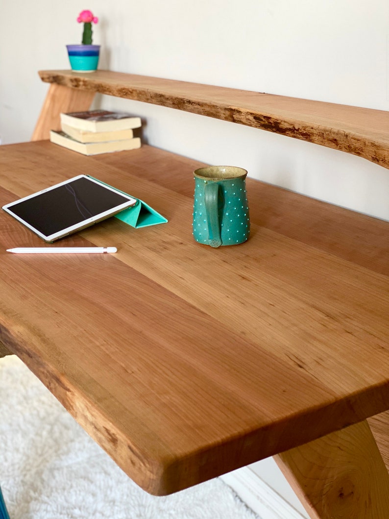 Pu&ograve; includere: Una scrivania in legno con bordo naturale e ripiano abbinato. Una tazza turchese con pois bianchi &egrave; appoggiata sulla scrivania, insieme a un tablet e uno stilo. Un piccolo cactus in un vaso blu e viola &egrave; sullo scaffale.