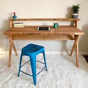 Pu&ograve; includere: Una scrivania in legno con finitura naturale, con un ripiano superiore e gambe angolate. Uno sgabello in metallo blu &egrave; posizionato di fronte. La scrivania contiene un tablet, una tazza e libri. Una piccola pianta in vaso e un cactus sono sulla scrivania.