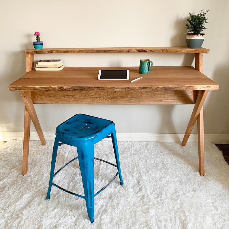 Pu&ograve; includere: Una scrivania in legno con finitura naturale, con un ripiano superiore e gambe angolate. Uno sgabello in metallo blu &egrave; posizionato di fronte. La scrivania contiene un tablet, una tazza e libri. Una piccola pianta in vaso e un cactus sono sulla scrivania.