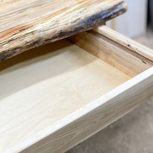 Pu&ograve; includere: Primo piano di una scrivania in legno con un cassetto aperto. La scrivania presenta un bordo naturale sulla parte superiore, con venature chiare. Il cassetto &egrave; anch'esso in legno chiaro, che mostra l'interno.