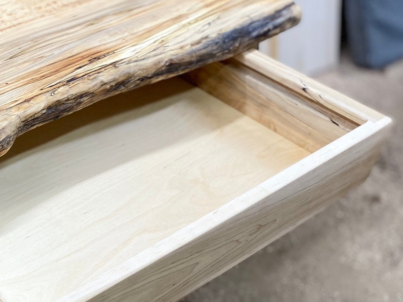 Pu&ograve; includere: Primo piano di una scrivania in legno con un cassetto aperto. La scrivania presenta un bordo naturale sulla parte superiore, con venature chiare. Il cassetto &egrave; anch'esso in legno chiaro, che mostra l'interno.
