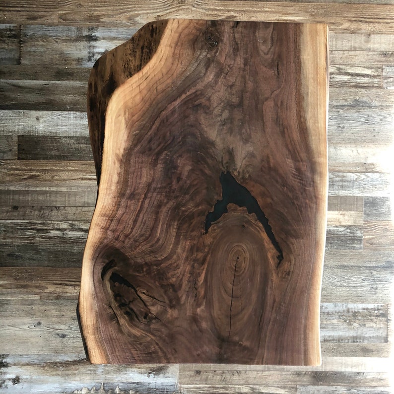Pu&ograve; includere: Una lastra rettangolare in legno di noce a bordo vivo con un motivo a venature naturali scure. La lastra ha una superficie liscia e lucida e un nodo scuro riempito di resina. La lastra di legno misura circa 76 cm di lunghezza e 51 cm di larghezza.