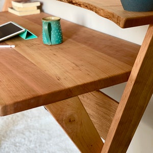 Pu&ograve; includere: Una scrivania in legno con un ripiano a bordo naturale. La scrivania ha una finitura marrone chiaro e un design a telaio a X. Una tazza turchese con pois bianchi &egrave; appoggiata sulla scrivania. Sono visibili anche un tablet e dei libri.