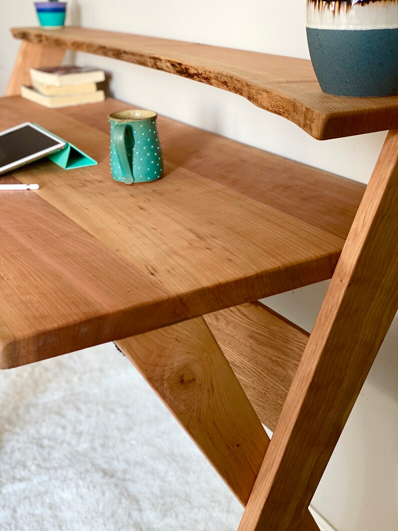 Pu&ograve; includere: Una scrivania in legno con un ripiano a bordo naturale. La scrivania ha una finitura marrone chiaro e un design a telaio a X. Una tazza turchese con pois bianchi &egrave; appoggiata sulla scrivania. Sono visibili anche un tablet e dei libri.