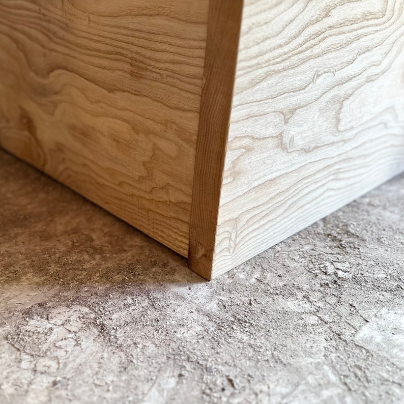 May include: Close-up of a light wood corner with a natural wood grain pattern. The corner is sitting on a light gray concrete surface.