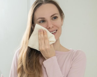 May include: A woman in a pink long-sleeved shirt is reflected in a mirror, holding a white cloth to her face.