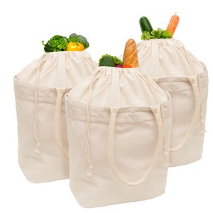 May include: Three off-white canvas tote bags filled with fresh produce. Each bag is open at the top, revealing broccoli, bell peppers, lettuce, cucumbers, carrots, and a baguette. The bags have drawstring closures and sturdy handles.
