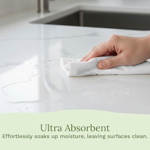 Op de afbeelding: Een witte, absorberende doek veegt water op een wit aanrechtblad. De tekst "Ultra Absorbent" wordt hieronder weergegeven, samen met de beschrijving "Effortlessly soaks up moisture, leaving surfaces clean."