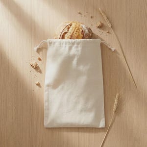 May include: A loaf of freshly baked bread peeks out of a cream-colored drawstring bag. The bread is golden brown with a textured crust. Wheat stalks and breadcrumbs are scattered on the light wooden surface, creating a rustic scene.