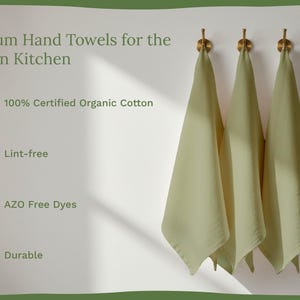 Op de afbeelding: Drie saliegroene handdoeken hangen aan gouden haken tegen een witte muur. De afbeelding bevat de tekst "Premium Hand Towels for the Modern Kitchen", "100% Certified Organic Cotton", "Lint-free", "AZO Free Dyes" en "Durable".