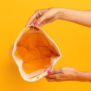 May include: A white canvas tote bag with a yellow lining. The bag is open and being held by a person's hands.