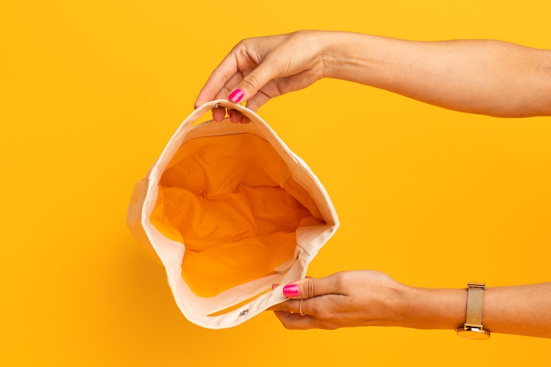 May include: A white canvas tote bag with a yellow lining. The bag is open and being held by a person's hands.