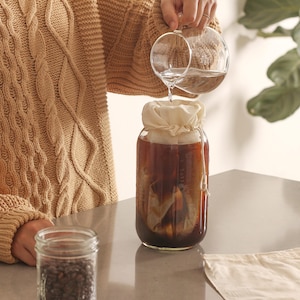 May include: A person pouring water into a glass jar with a white cloth filter. The jar is filled with a dark brown liquid. There is a bag of coffee beans in the foreground.