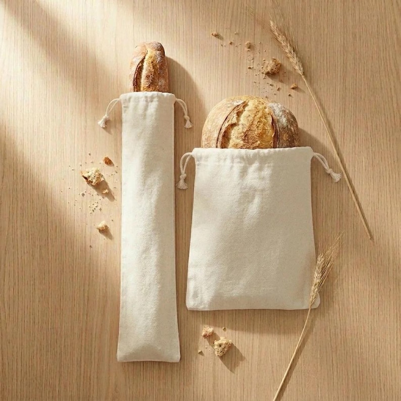 May include: Two off-white drawstring bread bags, one tall and narrow for a baguette, the other square for a round loaf. Both bags contain bread. Wheat stalks and breadcrumbs are scattered on the wooden surface.