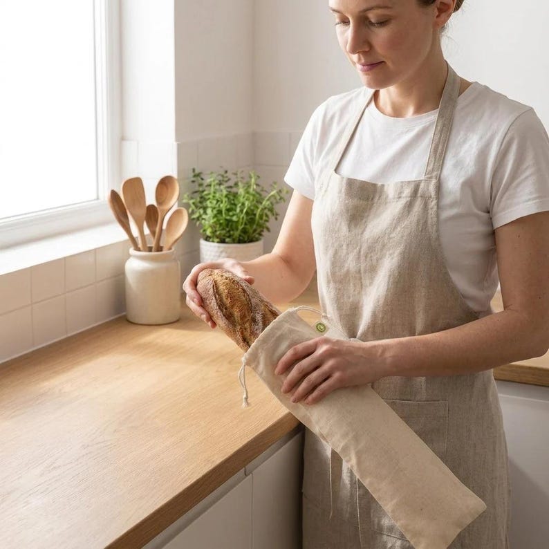 May include: A woman holding a fresh baguette, placing it into a beige linen bread bag. The bag has a drawstring closure and a small green tag. The kitchen features wooden spoons in a ceramic jar, a potted plant, and a light-colored countertop.