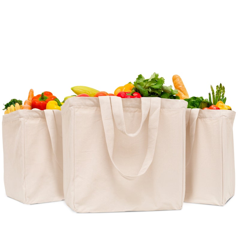May include: Three off-white canvas tote bags filled with fresh produce including green leafy vegetables, red bell peppers, yellow bananas, orange carrots, and a loaf of bread. Each bag features two sturdy handles and a rectangular base structure.