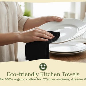 Op de afbeelding: Een zwarte keukendoek wordt gebruikt om een wit bord af te vegen. De afbeelding toont een stapel witte borden en de tekst "Eco-friendly Kitchen Towels" en "Made for 100% organic cotton for 'Cleaner Kitchens, Greener Planet.'"