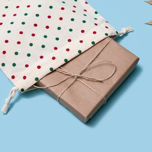 K&ouml;nnte beinhalten: Eine braune Geschenkbox, die mit Schnur umwickelt und mit einer Schleife gebunden ist, liegt in einer wei&szlig;en Stofftasche mit roten und gr&uuml;nen Punkten.