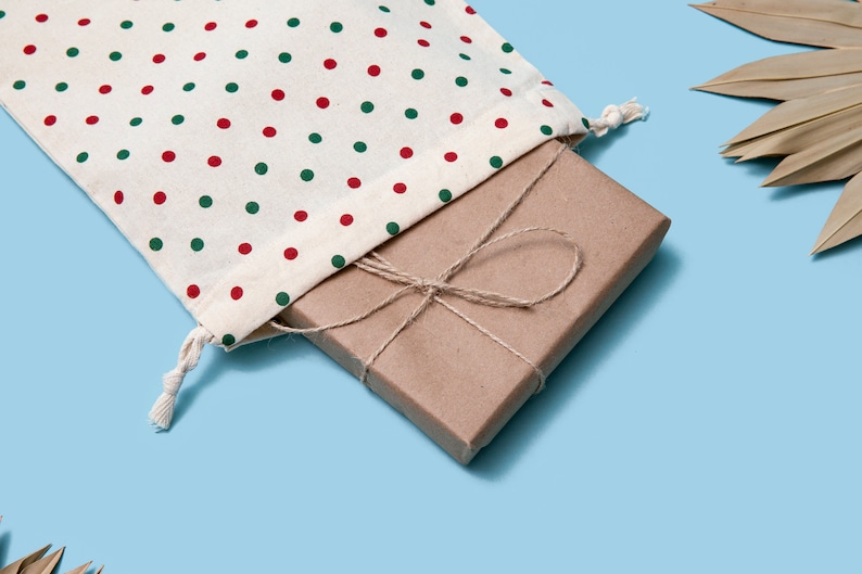 K&ouml;nnte beinhalten: Eine braune Geschenkbox, die mit Schnur umwickelt und mit einer Schleife gebunden ist, liegt in einer wei&szlig;en Stofftasche mit roten und gr&uuml;nen Punkten.