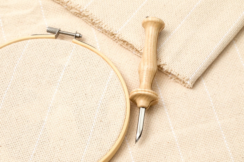 May include: A wooden embroidery needle tool and a round embroidery hoop on a beige fabric with white stripes.