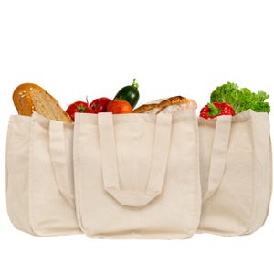May include: Three off-white canvas tote bags filled with fresh groceries. The bags contain a loaf of bread, red tomatoes, a green cucumber, and a head of green lettuce. The bags feature sturdy fabric handles and a simple, rectangular construction.