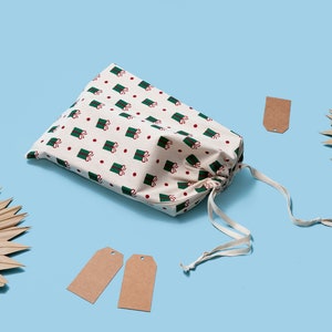 K&ouml;nnte beinhalten: Eine wei&szlig;e Stofftasche mit einem gr&uuml;nen und roten Muster aus verpackten Geschenken. Die Tasche ist mit einem wei&szlig;en Kordelzug gebunden und liegt auf einer hellblauen Oberfl&auml;che. Auf der Oberfl&auml;che befinden sich drei braune Geschenkanh&auml;nger aus Papier.