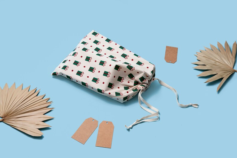 K&ouml;nnte beinhalten: Eine wei&szlig;e Stofftasche mit einem gr&uuml;nen und roten Muster aus verpackten Geschenken. Die Tasche ist mit einem wei&szlig;en Kordelzug gebunden und liegt auf einer hellblauen Oberfl&auml;che. Auf der Oberfl&auml;che befinden sich drei braune Geschenkanh&auml;nger aus Papier.