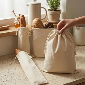 May include: Beige canvas bread bags with drawstring closures. Several bags of different sizes hold various types of bread. A hand is holding one of the bags. A baguette is in a long, narrow bag.