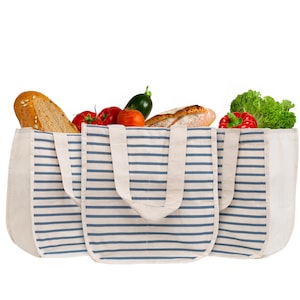 May include: A set of three rectangular tote bags made of off-white fabric with horizontal navy blue stripes. Each bag features two sturdy fabric handles and is filled with various fresh groceries, including bread, tomatoes, and green lettuce.