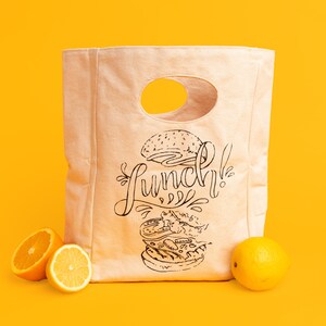May include: A beige canvas lunch bag with a black and white illustration of a hamburger and the word "Lunch!" printed on it. Two orange halves are in front of the bag.