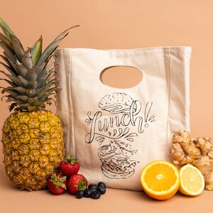 May include: A white canvas lunch bag with a black and white illustration of a burger and the word "Lunch!" printed on it. The bag is surrounded by a pineapple, strawberries, blueberries, oranges, and ginger.