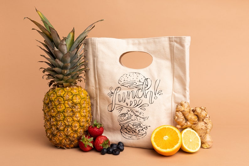 May include: A white canvas lunch bag with a black and white illustration of a burger and the word "Lunch!" printed on it. The bag is surrounded by a pineapple, strawberries, blueberries, oranges, and ginger.