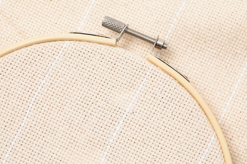 May include: A close-up of a wooden embroidery hoop with a beige fabric stretched across it. The hoop is held together with a metal screw.