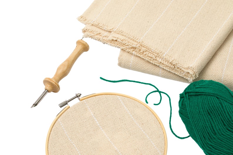 May include: A set of embroidery supplies including a wooden embroidery hoop, a skein of green yarn, a needle threader, and a piece of beige Aida cloth.