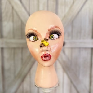 May include: A white mannequin head with painted-on eyes, lips, and eyebrows. A small yellow and black bee figurine is attached to the mannequin's nose.