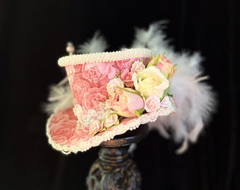 Puede incluir: Un sombrero de copa rosa con un borde de encaje blanco y un diseño floral. El sombrero está decorado con flores rosas y blancas y una pluma blanca.