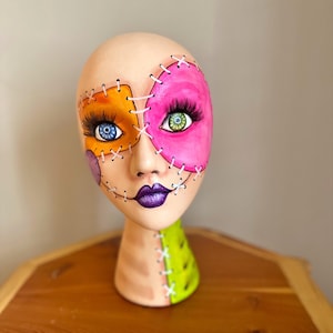 May include: A painted mannequin head with a patchwork design. The face is divided into orange and pink sections, stitched together with white thread. The eyes are blue and green, with long lashes. The lips are purple, and the neck is green.