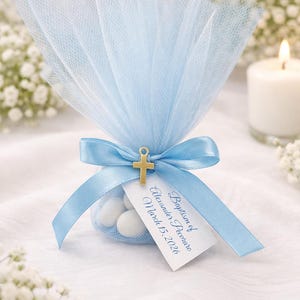 Puede incluir: Una bolsita de tul azul claro atada con una cinta a juego y un dije de cruz dorada. La bolsa contiene caramelos blancos y una etiqueta que dice "Baptismo de Alexander Pecoraro 15 de marzo de 2026."